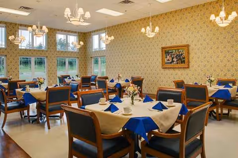 An empty senior living dining room with round tables, blue napkins, flowers, and chandeliers.