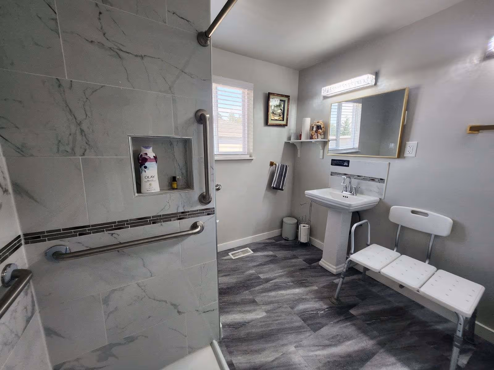 A modern bathroom with gray marble-patterned tiles in the shower area, equipped with grab bars for accessibility. The bathroom has a white pedestal sink with a mirror above it, a white bench with three seats, a small window with blinds, a towel hanging on a wall hook, and a shelf with decorative items. The floor has gray wood-like vinyl flooring.
