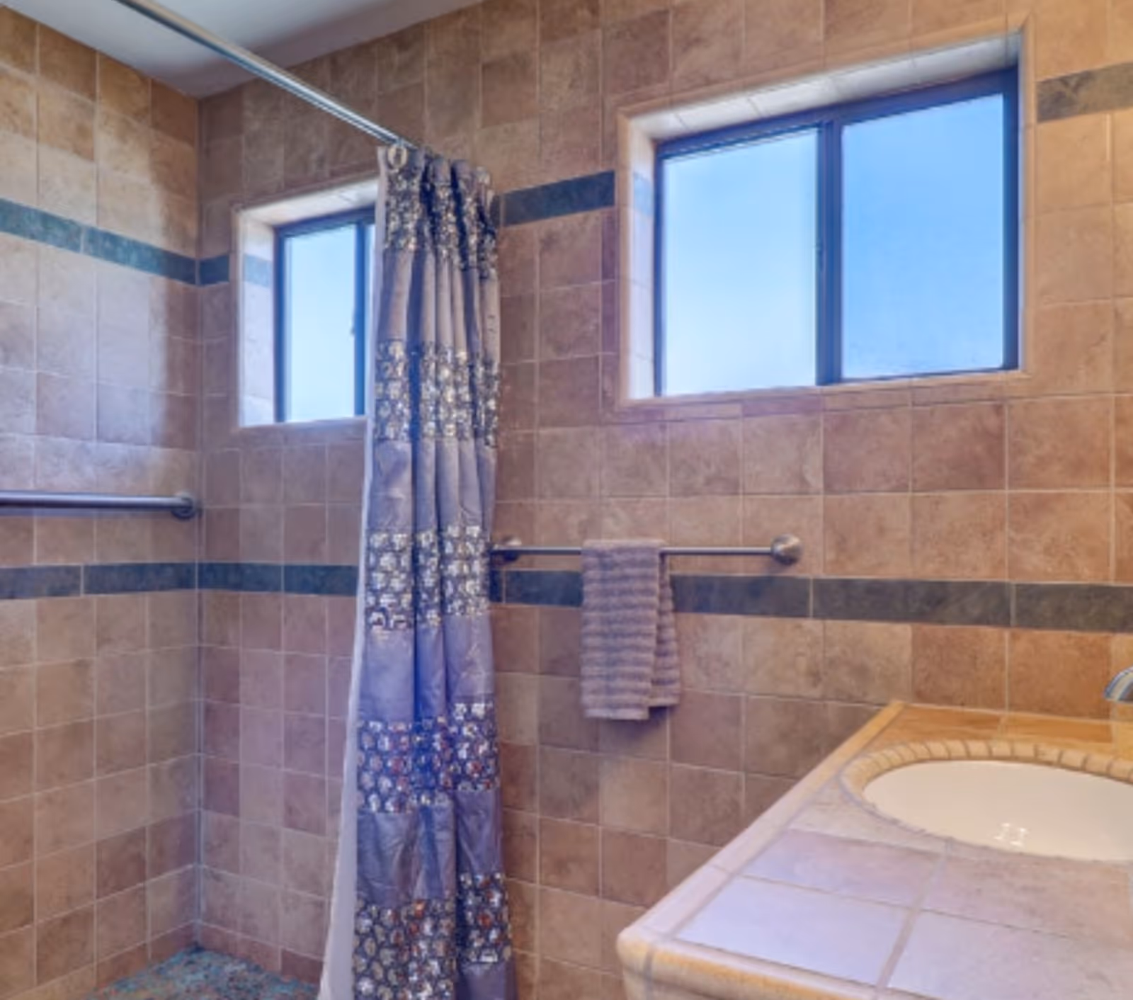 A bathroom with tiled walls and floor, featuring a shower area with a decorative shower curtain, a window with frosted glass, a towel hanging on a metal bar, and a tiled countertop with a built-in sink.