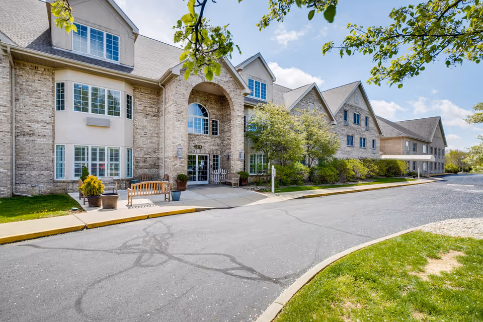Exterior view of a large brick assisted living facility building with multiple windows and a main entrance featuring an arched design. There are benches and potted plants near the entrance, a paved driveway, and green trees and grass surrounding the building under a partly cloudy sky.