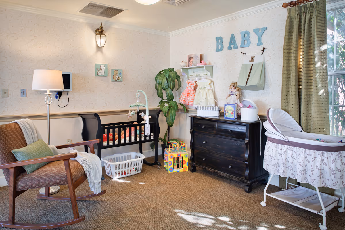 Bright nursery-style sitting room with a rocking chair, crib, dresser, bassinet, toys and wall letters spelling 'BABY'.