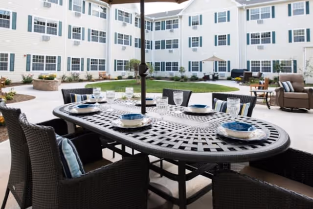 Outdoor courtyard area with a large black metal table set with plates and glasses, surrounded by wicker chairs with cushions. The courtyard is enclosed by a three-story white building with many windows. There are additional seating areas with chairs and tables, green grass, and landscaping.