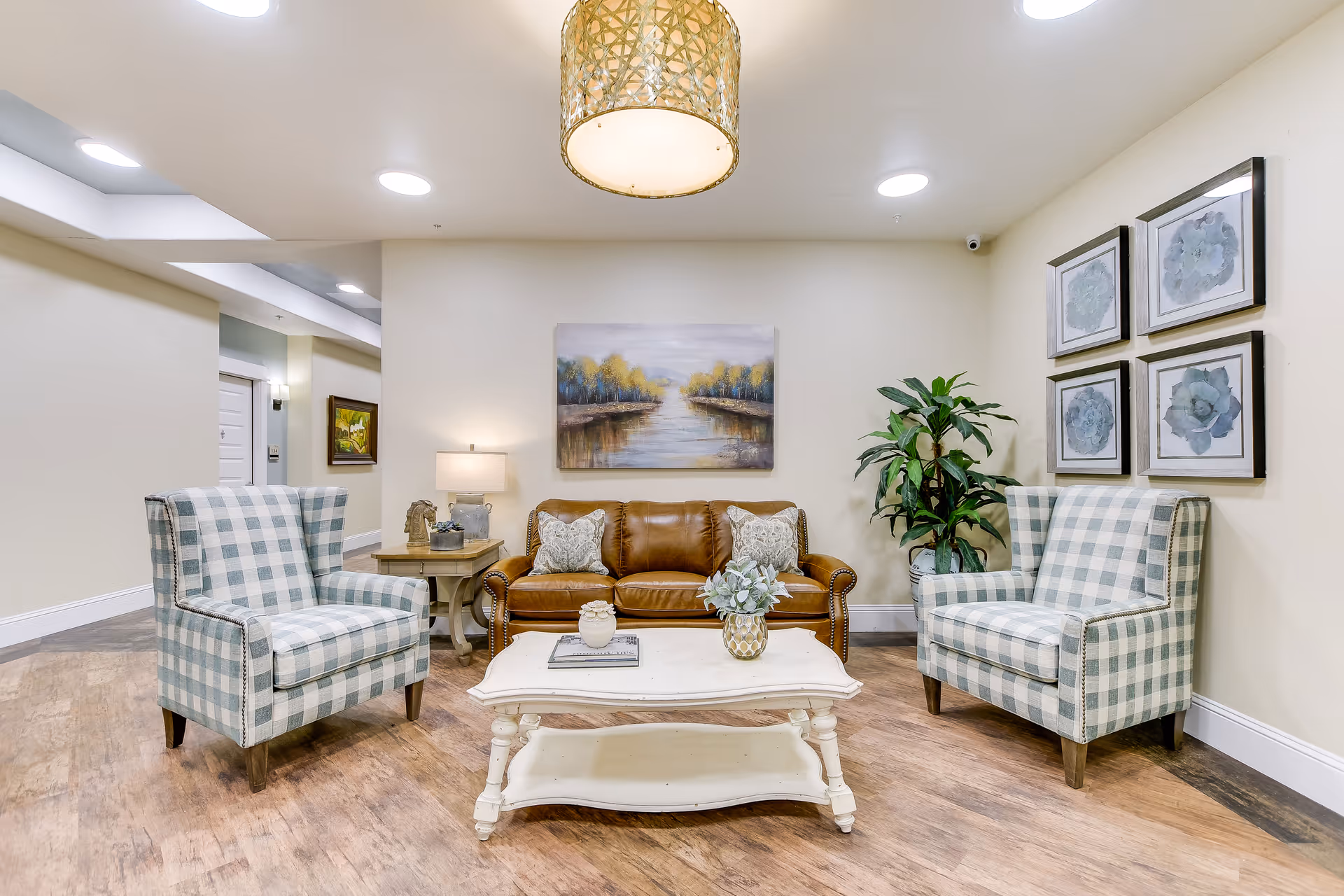A cozy sitting area in a senior living facility featuring a brown leather sofa with two patterned pillows, two plaid armchairs, a white wooden coffee table with decorative items, a side table with a lamp and a small sculpture, a large potted plant, and framed artwork on the walls. The floor is wood, and the ceiling has recessed lighting and a woven pendant light.