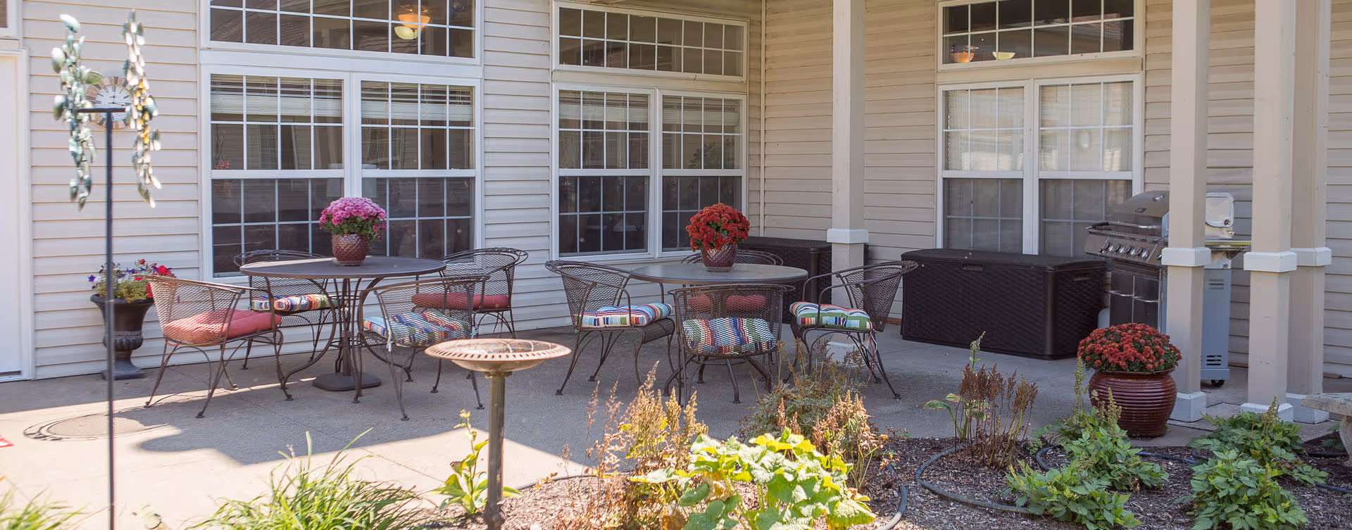Outdoor patio area with metal tables and chairs featuring colorful cushions. There are potted plants with flowers on the tables and around the patio. The patio is adjacent to a building with large windows and beige siding. A grill and storage containers are visible in the corner.