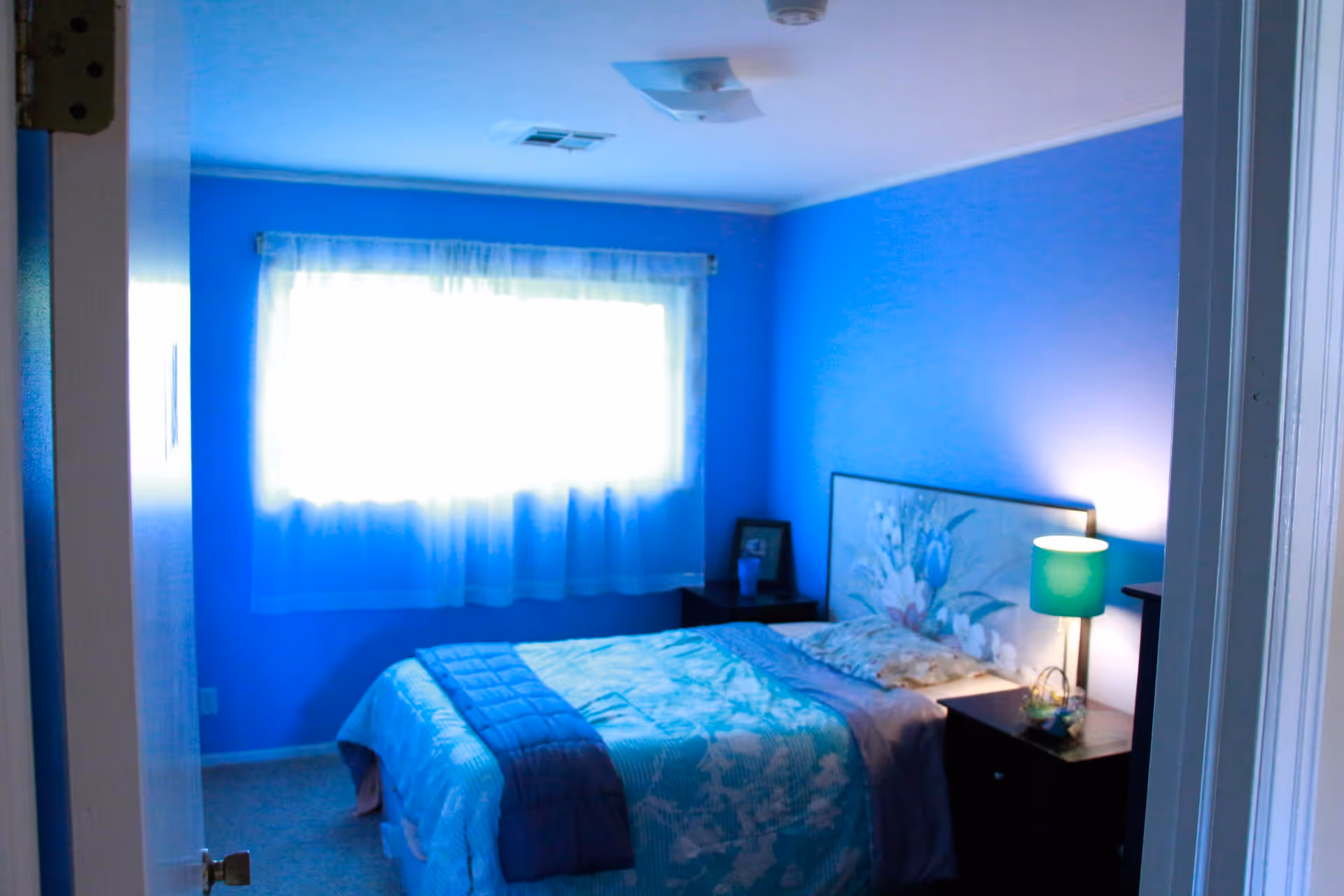 A bedroom with bright blue walls and a window covered with sheer white curtains. The bed has a floral headboard and is made with a blue and white patterned comforter and a dark blue throw blanket. There are two dark wooden nightstands on either side of the bed, one with a green lamp and a small basket, and the other with a framed photo and a cup.