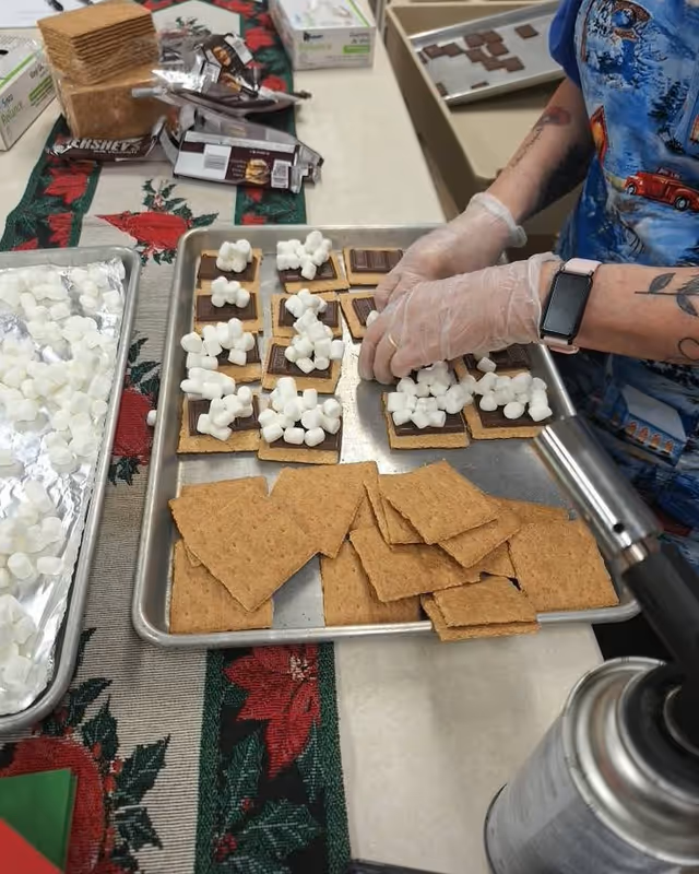 A close-up of gloved hands assembling marshmallow-topped crackers on a metal tray in a kitchen prep area.