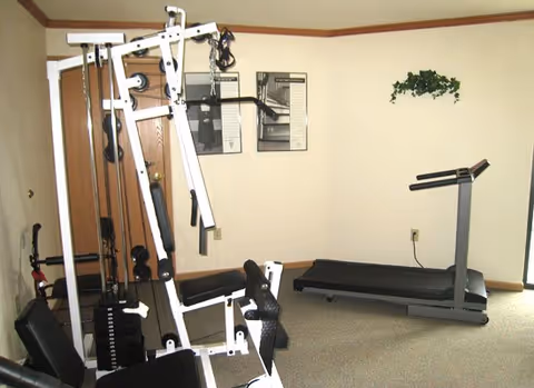 A small fitness room with exercise equipment including a multi-station weight machine and a treadmill. The room has beige walls with a small green plant decoration and two framed posters on the wall.