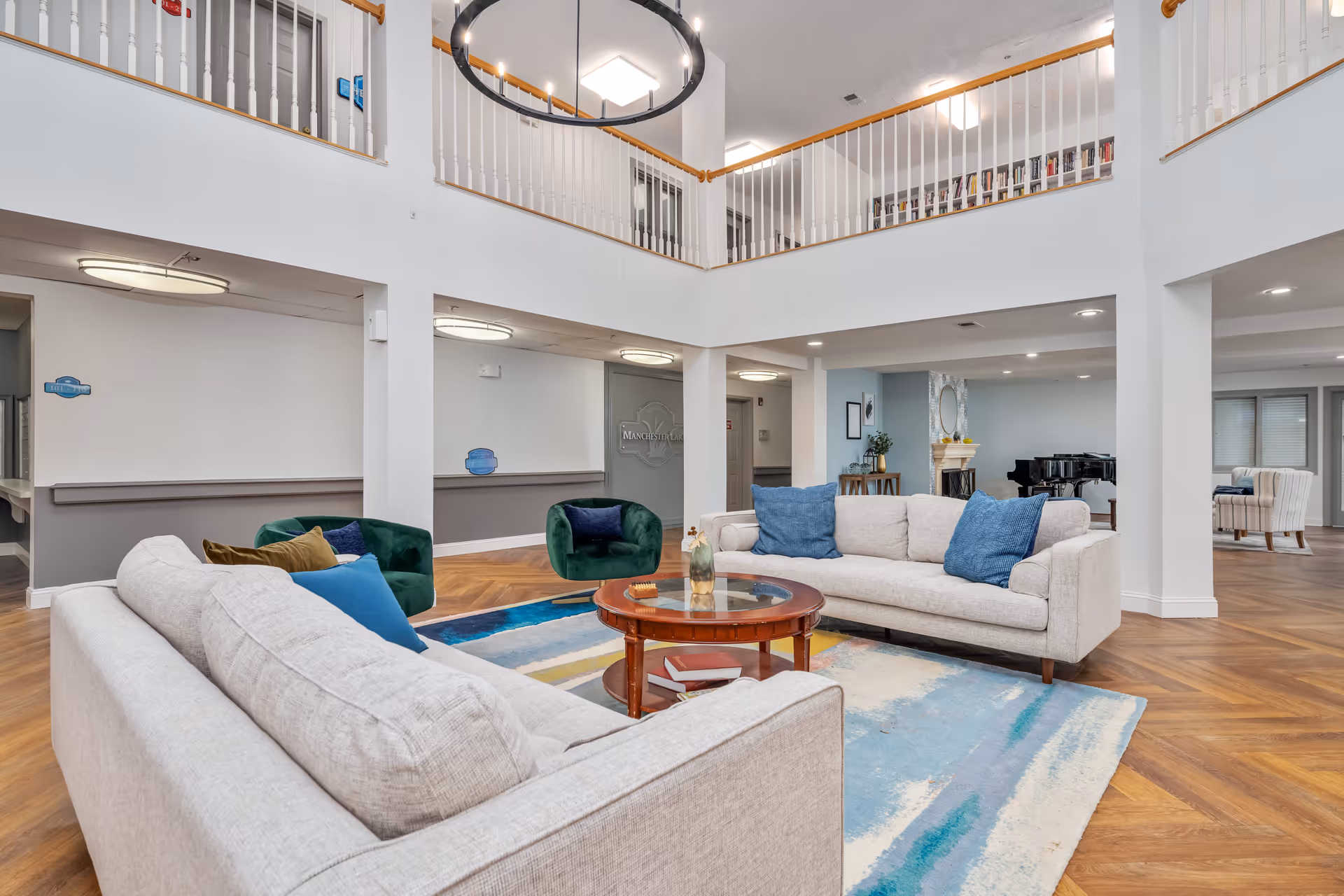 A spacious senior living facility common area with two beige sofas adorned with blue and brown pillows, a round wooden coffee table with decorative items, two green armchairs, and a colorful area rug. The room features high ceilings with a modern circular chandelier, white walls, wooden flooring, and a second-floor balcony railing overlooking the space. In the background, there is a piano and additional seating areas.