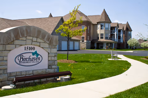 Exterior view of Birchaven Retirement Village featuring a stone sign with the facility name and address 15100, a curved sidewalk, green lawn, young trees, and a multi-story building with peaked roofs and balconies under a clear blue sky.