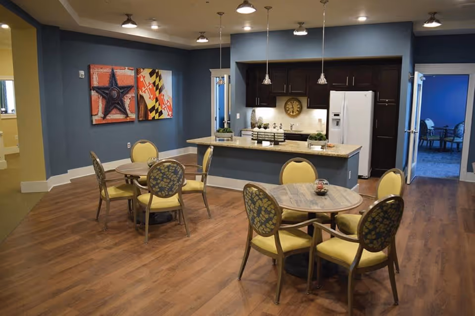 Interior view of a senior living facility dining area with two round wooden tables, each surrounded by four yellow cushioned chairs with patterned backs. The room features wood flooring, blue walls, and a kitchen area with dark cabinets, a white refrigerator, a clock on the wall, and pendant lights hanging above a granite countertop. Two colorful wall art pieces are displayed on the left wall, and an open doorway leads to another room with blue walls and additional seating.