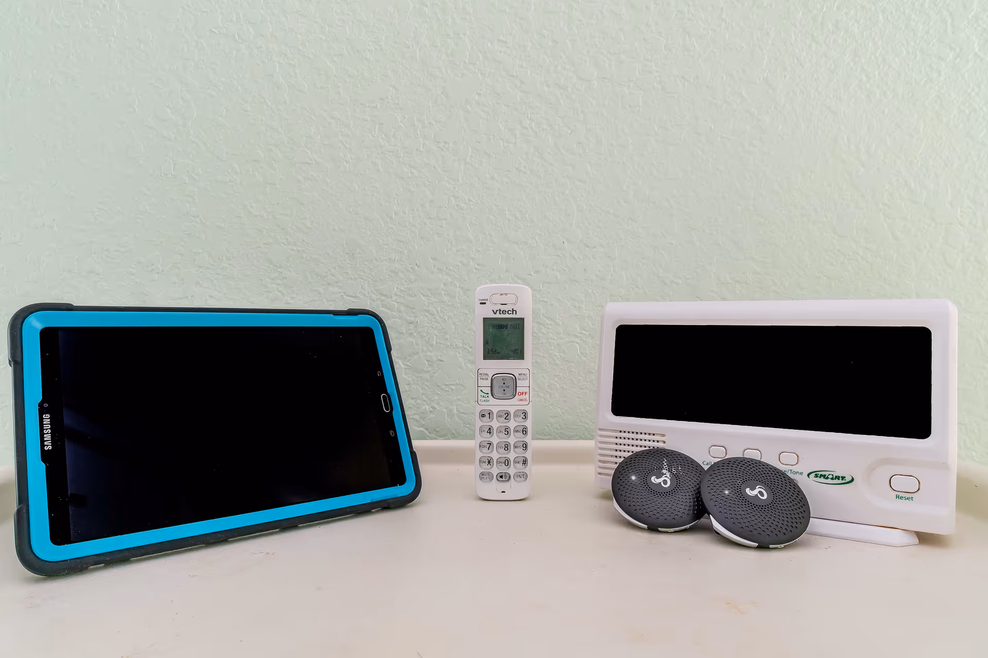 A Samsung tablet with a blue and black protective case, a white VTech cordless phone, and a white electronic device with two round black speakers placed on a light-colored surface against a pale green textured wall.
