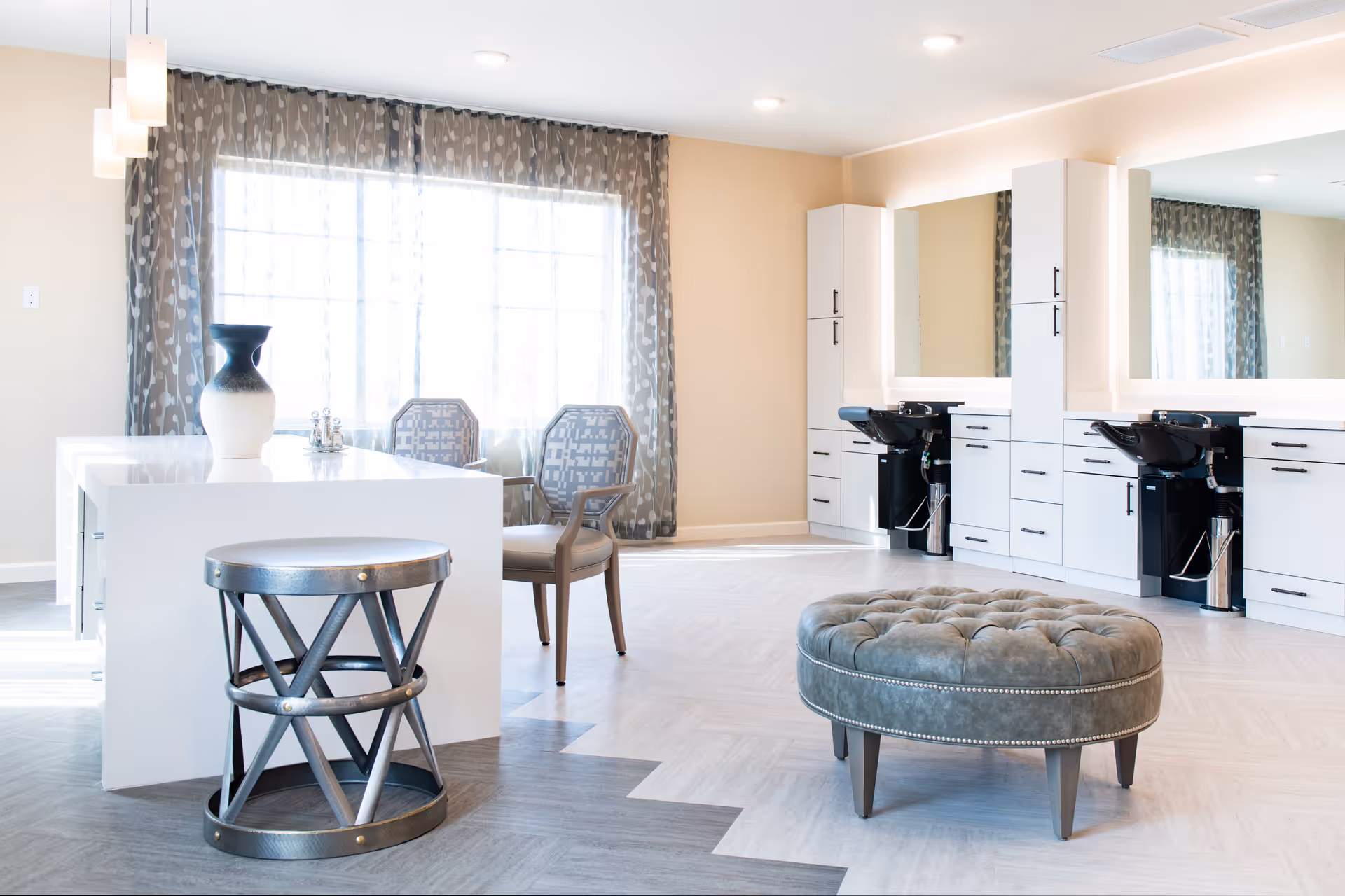 Bright and modern salon area with two black hair washing stations, large mirrors, white cabinetry, a white island table with a decorative vase, two patterned chairs near a large window with sheer curtains, a round tufted ottoman, and a metal stool on a light and dark gray patterned floor.