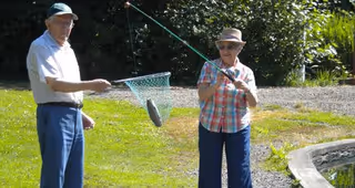 An elderly man and woman fishing outdoors. The man is holding a fishing net with a fish caught in it, while the woman is holding a fishing rod. They are standing on grass near a small pond with trees and bushes in the background.