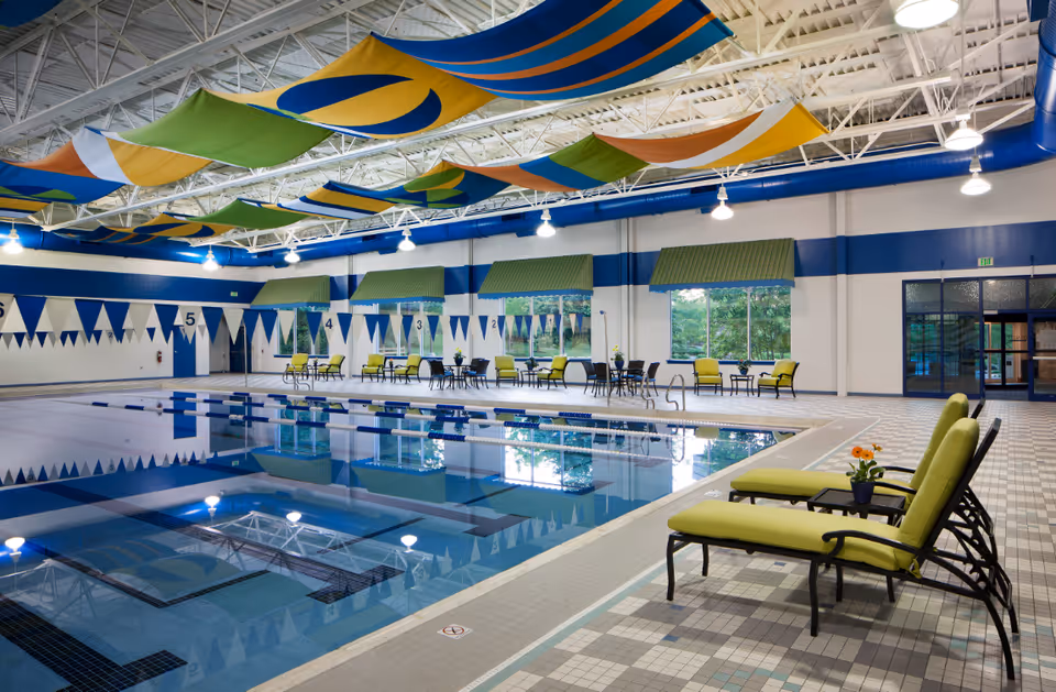 Indoor community swimming pool with lanes, lounge chairs, tables, and colorful fabric banners hanging from the ceiling.