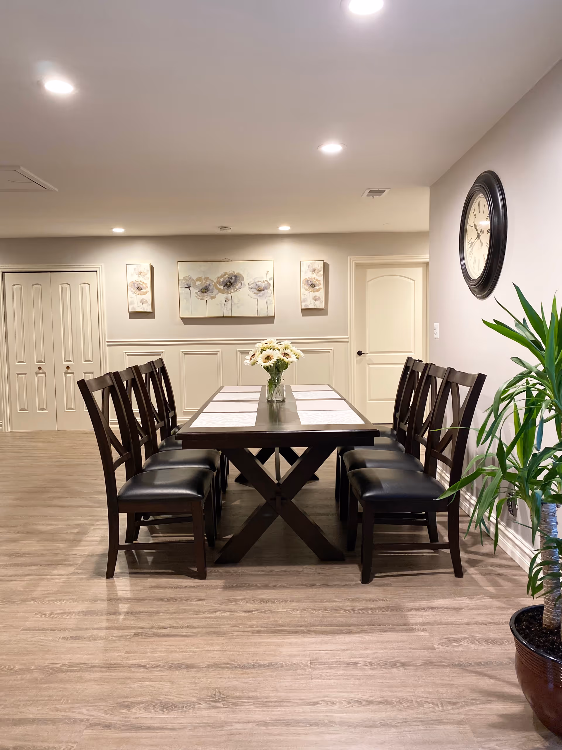 A long wooden dining table with eight chairs and a vase of flowers in a bright room with wall art and a large clock on the wall.
