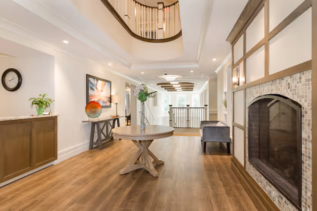 A bright and spacious interior hallway with wooden flooring, a round wooden table with a vase of flowers in the center, a fireplace with a tiled surround on the right, a gray armchair, a console table with decorative items and a wall clock on the left, and a staircase with a curved railing leading to an upper floor.