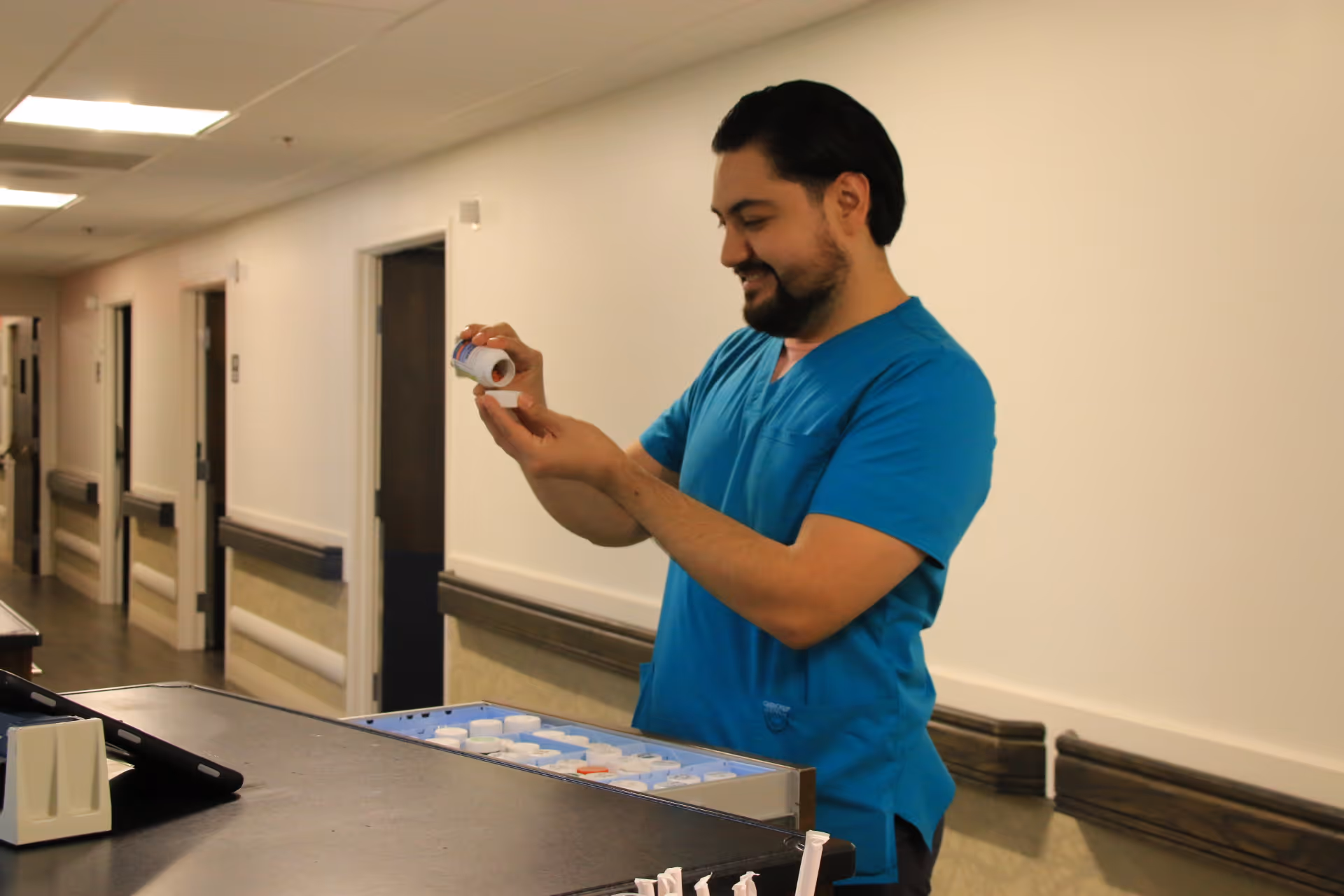 A healthcare worker in blue scrubs standing at a medication cart in a hallway, holding and examining a pill bottle with a smile.