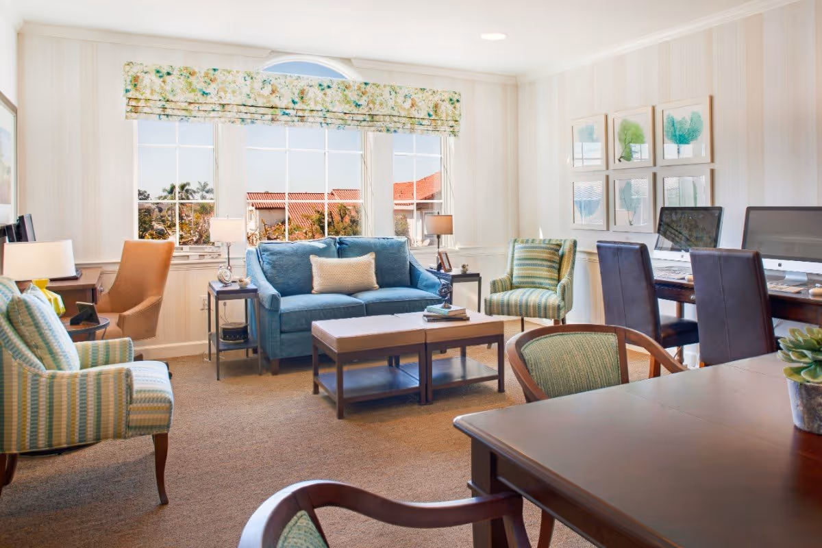 A bright and cozy living room with a blue sofa, two striped armchairs, a brown armchair, two side tables with lamps, a coffee table, and a large window with floral valance. There are framed botanical prints on the wall and a desk area with two computers and dark chairs.