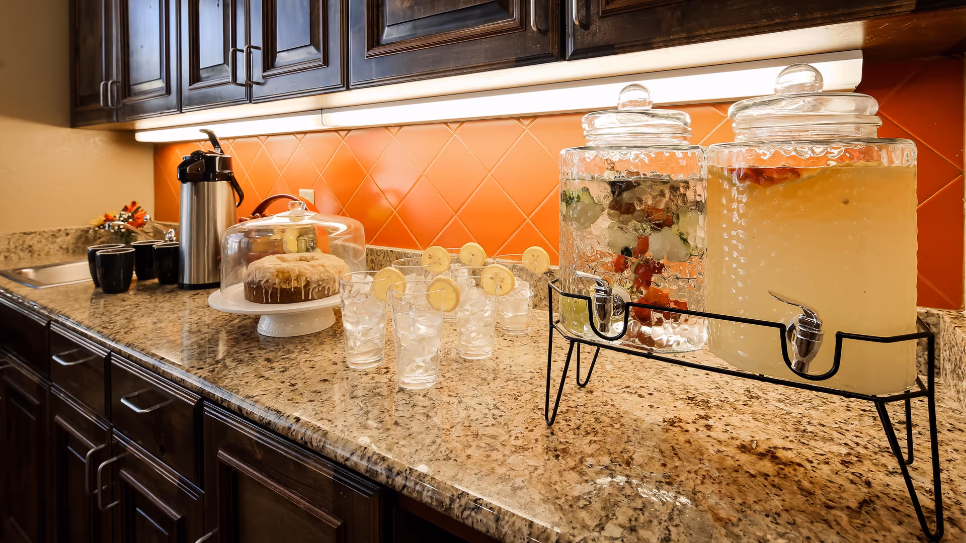 Granite countertop buffet with beverage dispensers, glasses garnished with lemon slices, a covered cake, a coffee urn, and dark cabinets against an orange tiled backsplash.