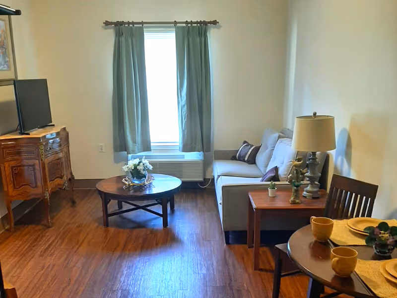 A cozy living room with wooden flooring, a gray sofa with cushions, a wooden coffee table with a flower arrangement, a side table with a lamp and small decorative items, a wooden dining table with two yellow cups and placemats, and a wooden cabinet with a TV on top. A window with light blue curtains is in the background.