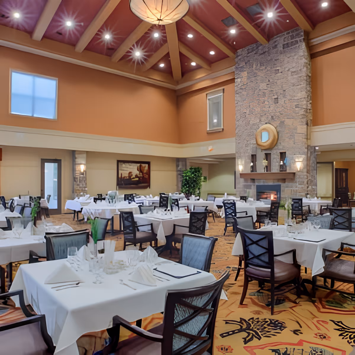 Spacious dining room with multiple tables covered in white tablecloths, set with glassware and napkins. The room features a high ceiling with wooden beams, a large stone fireplace with a decorative mirror above it, and warm-toned walls. There are several chairs around each table and a patterned carpet on the floor.