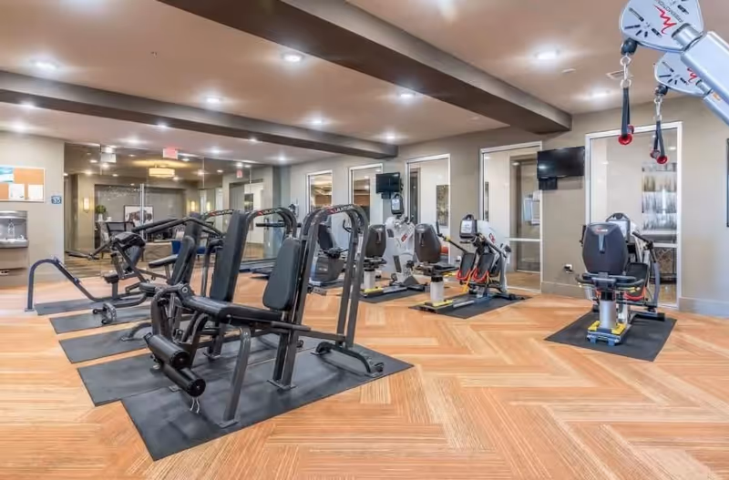 A spacious fitness room with various exercise machines including weight machines and recumbent bikes, wooden flooring, bright ceiling lights, and large mirrors on one wall.