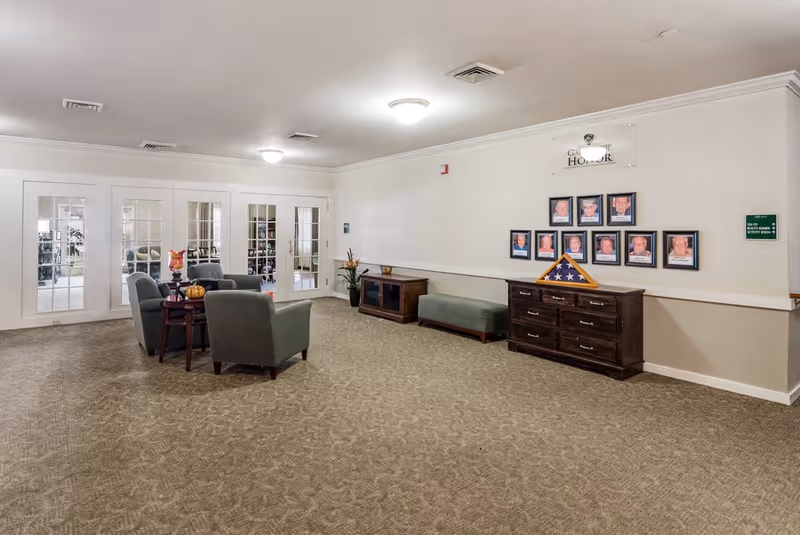 A spacious, carpeted common area with several armchairs arranged around a small table. The room has white walls with a wooden sideboard and a bench along one wall. Above the sideboard is a display of framed photos and a folded American flag in a triangular case. There are double glass doors leading to another room with bookshelves visible inside.