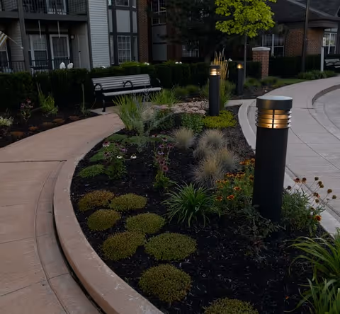 Curved landscaped walkway with planted beds, illuminated bollard lights, a bench, and an apartment building in the background.