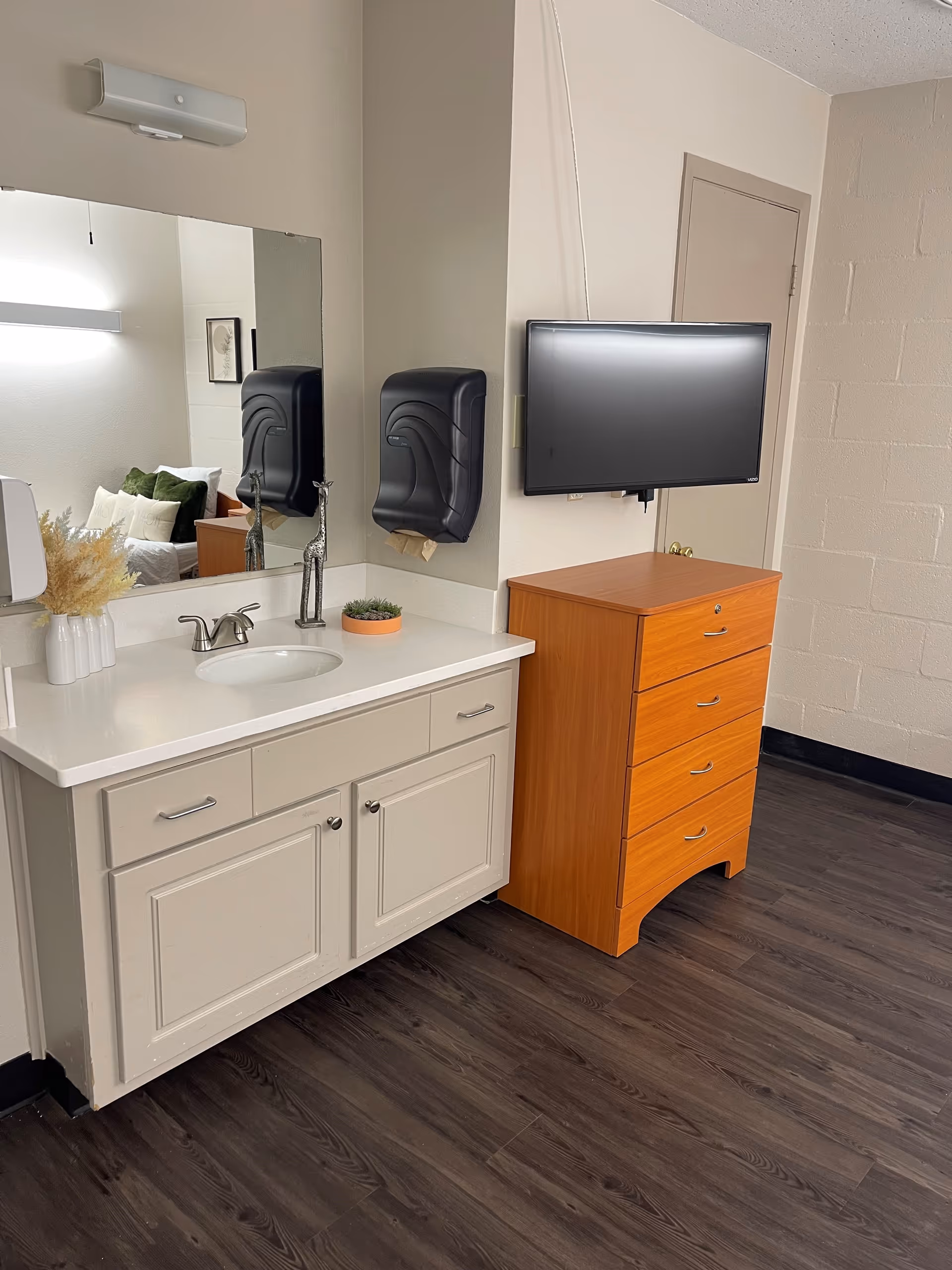 A room corner featuring a white vanity with a sink, a large mirror above it, and a mounted black paper towel dispenser. Next to the vanity is a wooden chest of drawers with a flat-screen TV mounted on the wall above it. The floor is dark wood, and a door is visible in the background.