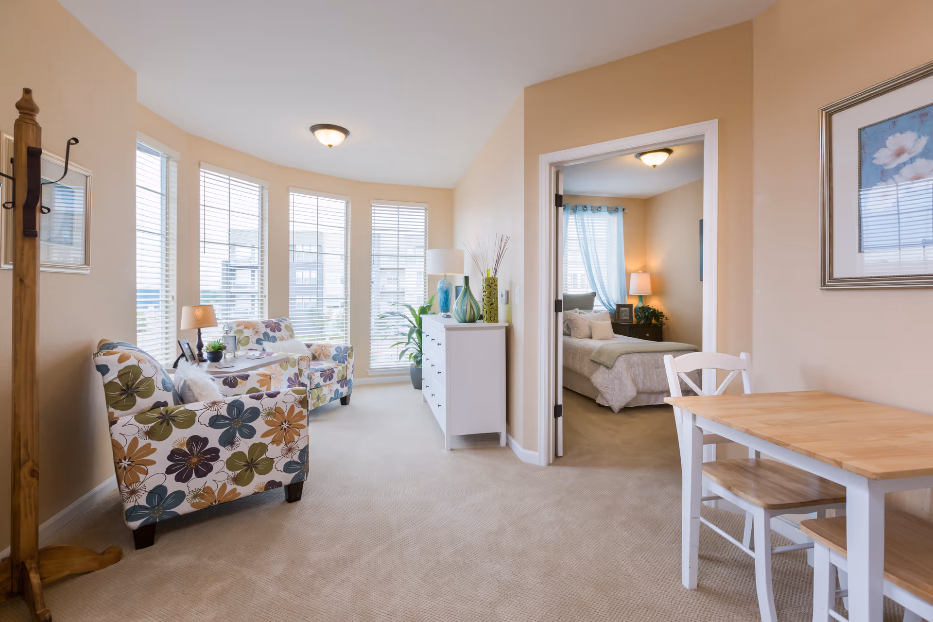 Bright living area with floral armchairs by bay windows, a small dining table, a white dresser, and a view into a bedroom.
