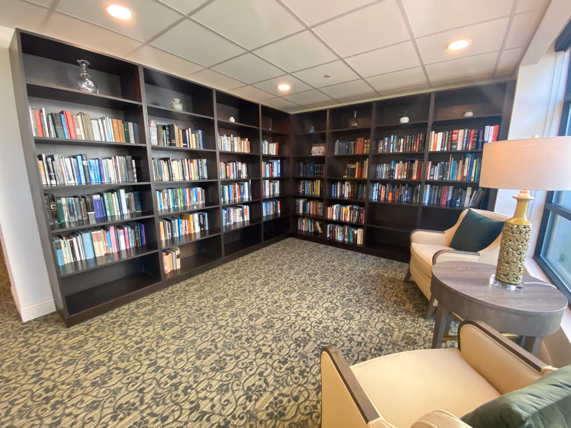 A cozy library corner in a senior living facility with dark wooden bookshelves filled with books lining two walls. Two beige armchairs with green cushions are positioned near a round wooden side table with a decorative lamp. The room has a patterned carpet and a window letting in natural light.