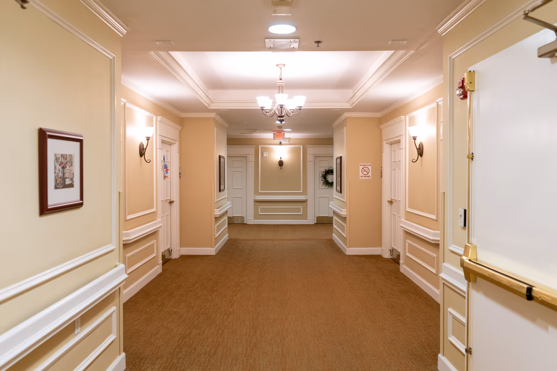 A well-lit hallway in a senior living facility with beige walls, white trim, and brown carpet. The hallway features multiple white doors on both sides, wall sconces providing light, framed artwork on the walls, and a chandelier hanging from the ceiling. There is an exit sign and a no smoking sign visible at the end of the hallway.