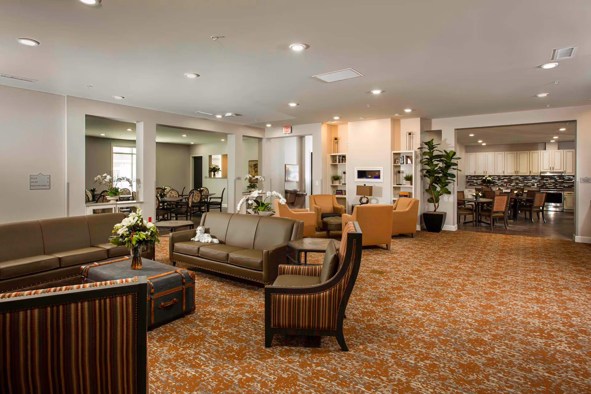 Bright, spacious communal lounge with sofas, armchairs, tables, plants, and a view into a dining area and kitchen.