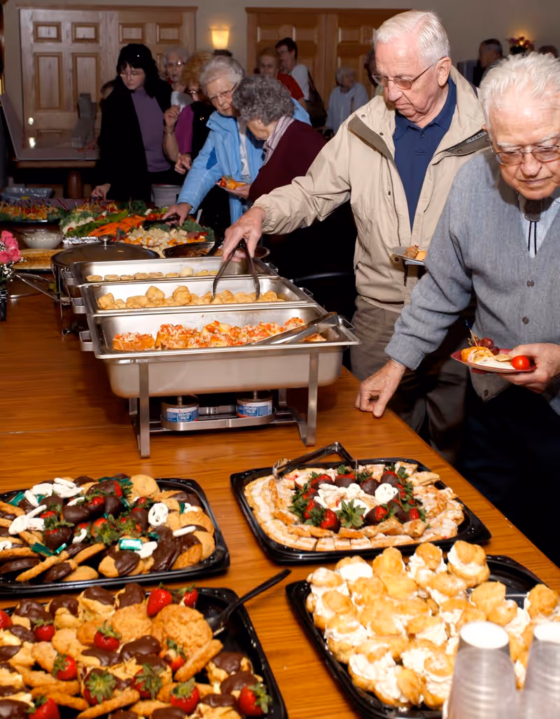 A group of elderly people serving themselves food from a buffet table filled with trays of various appetizers and desserts in a dining area.