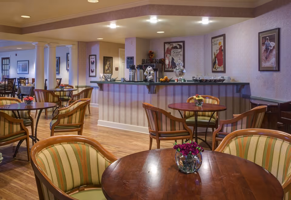 Cozy bistro-style dining room with round wooden tables, striped upholstered chairs, and a serving counter against a wallpapered wall.