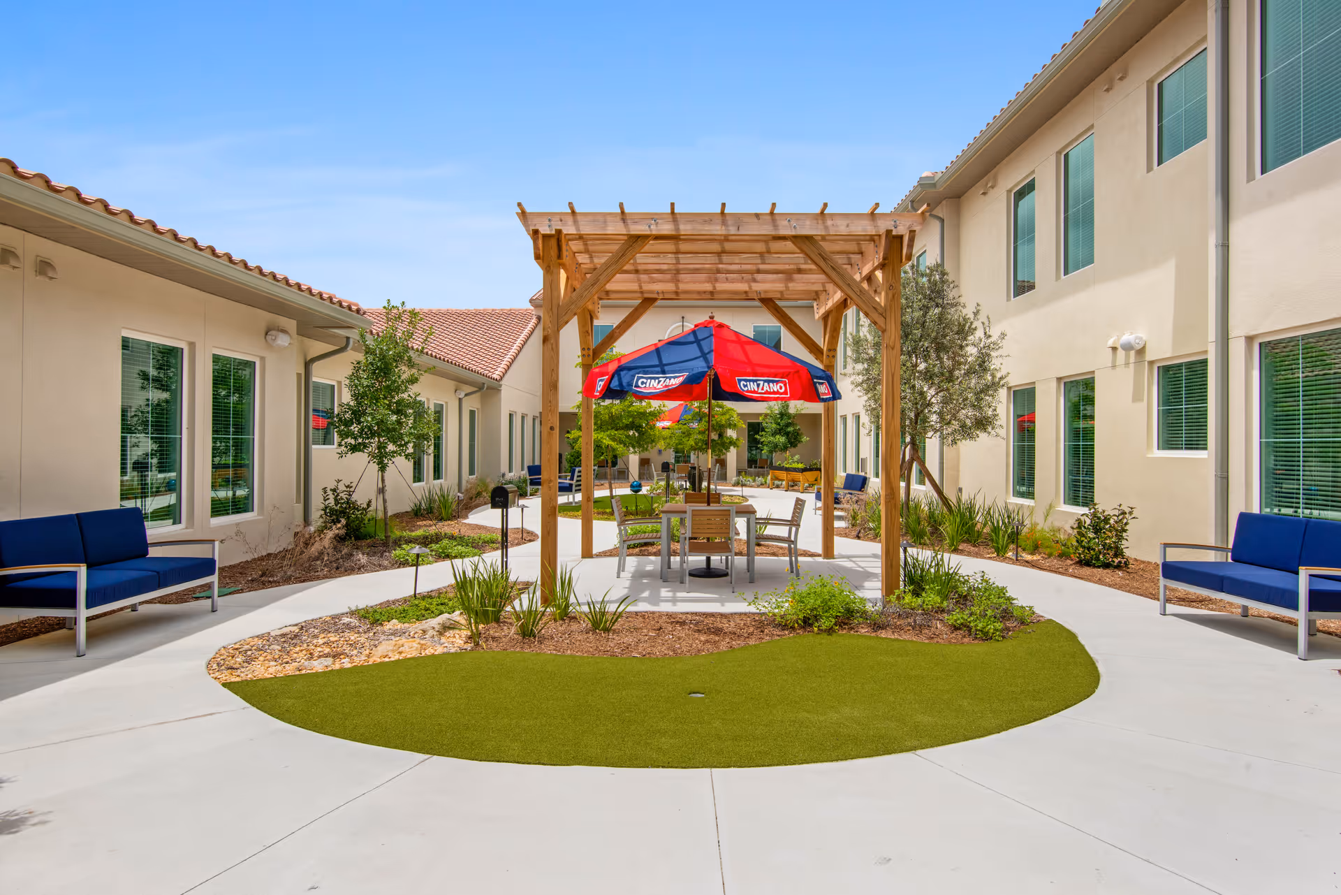 Outdoor courtyard area at Tuscan Gardens Of Delray Beach featuring a wooden pergola with a red and blue Cinzano umbrella over a table and chairs. The courtyard is surrounded by beige buildings with multiple windows and blue cushioned benches along the walkway. There are small landscaped garden beds with plants and trees, and a clear blue sky overhead.