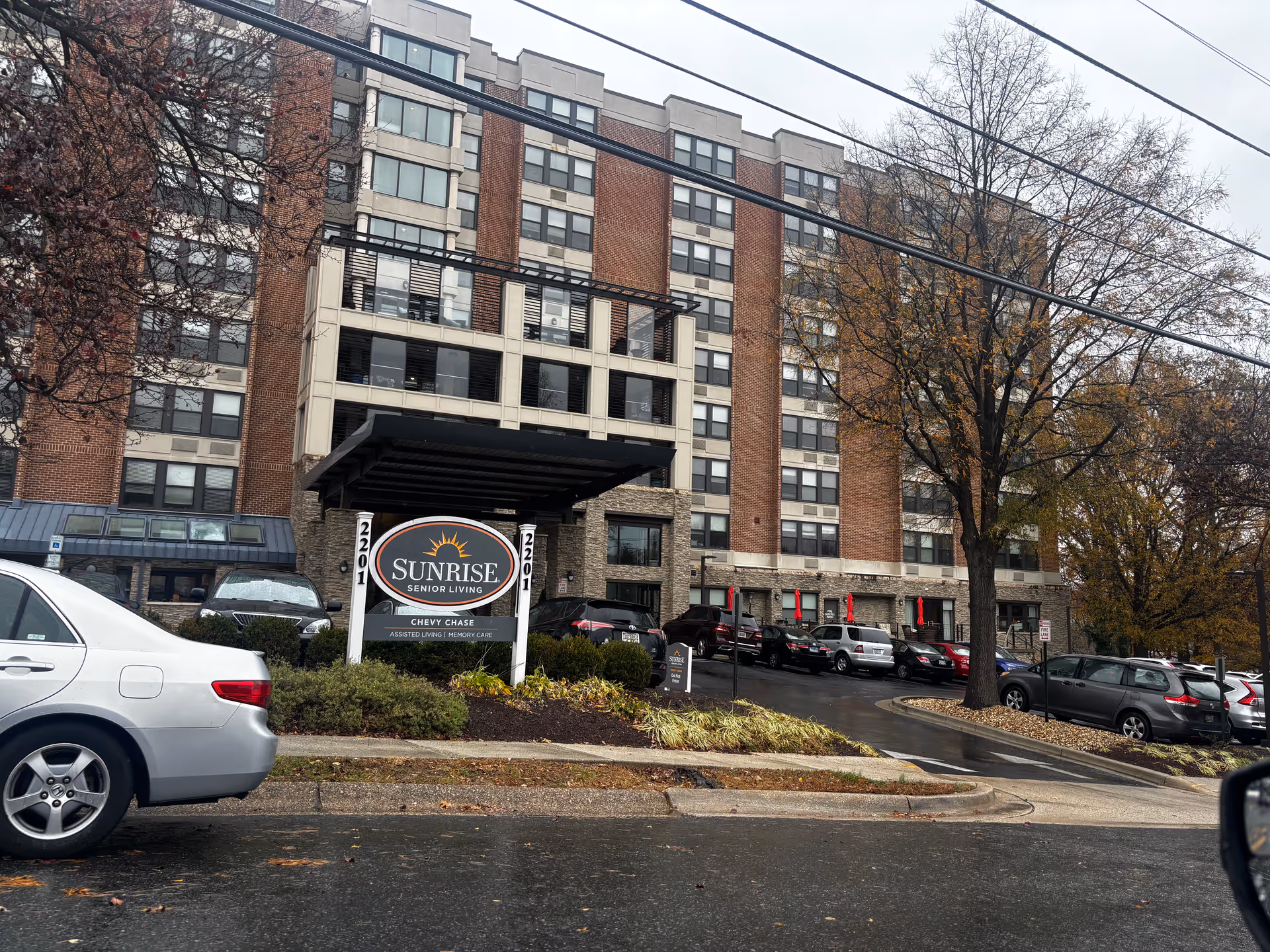 Front exterior of the Sunrise of Chevy Chase senior living building with its entrance sign and parked cars.