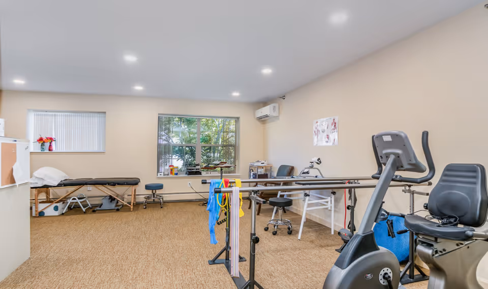 A physical therapy or rehabilitation room with exercise equipment including a stationary bike, parallel bars with resistance bands, a treatment table, stools, chairs, and anatomical charts on the wall. The room has beige walls, a large window showing greenery outside, and recessed ceiling lights.