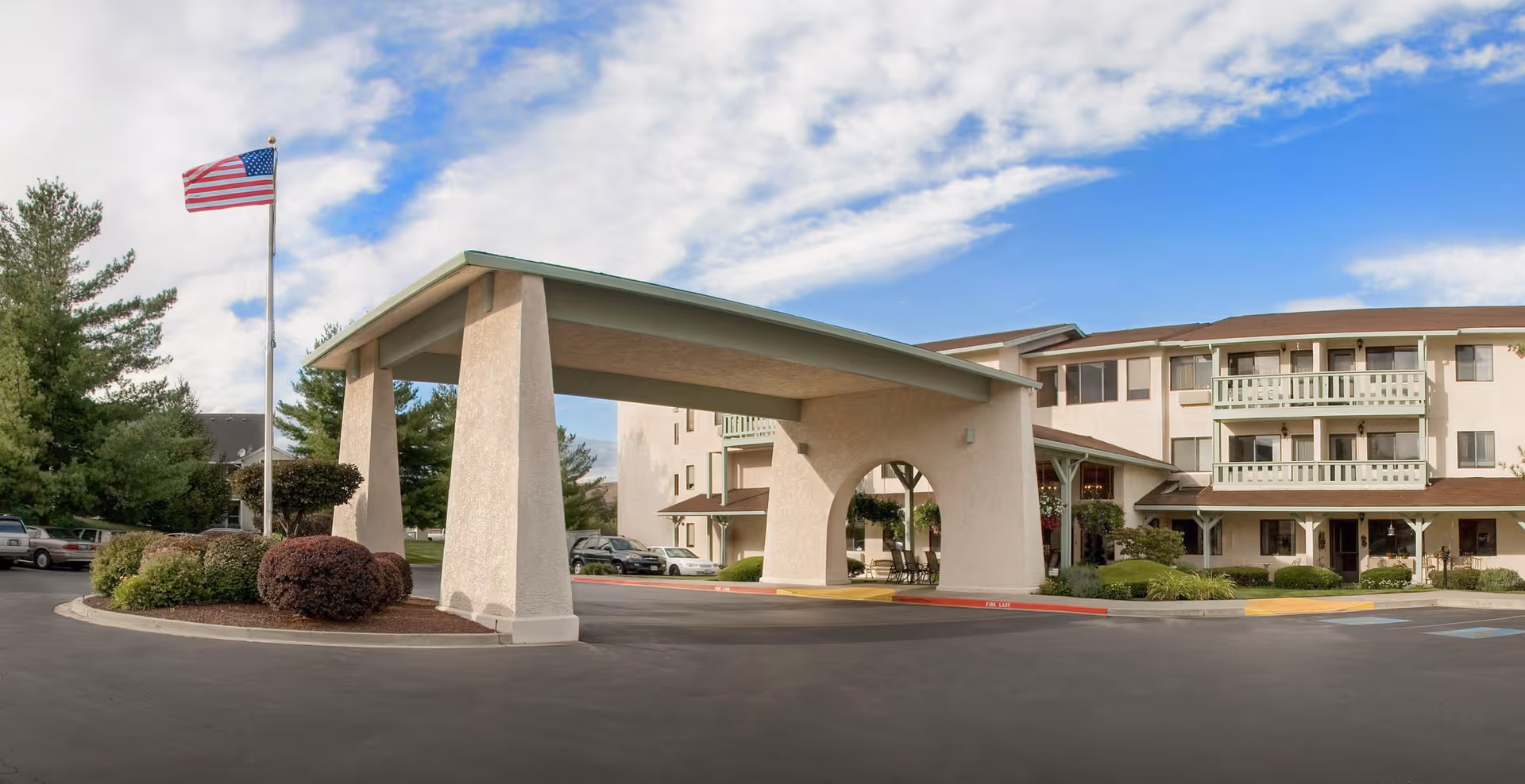 Exterior view of Solista Yakima senior living facility showing a covered entrance with large pillars, an American flag on a flagpole, landscaped bushes, and a multi-story building with balconies under a partly cloudy sky.
