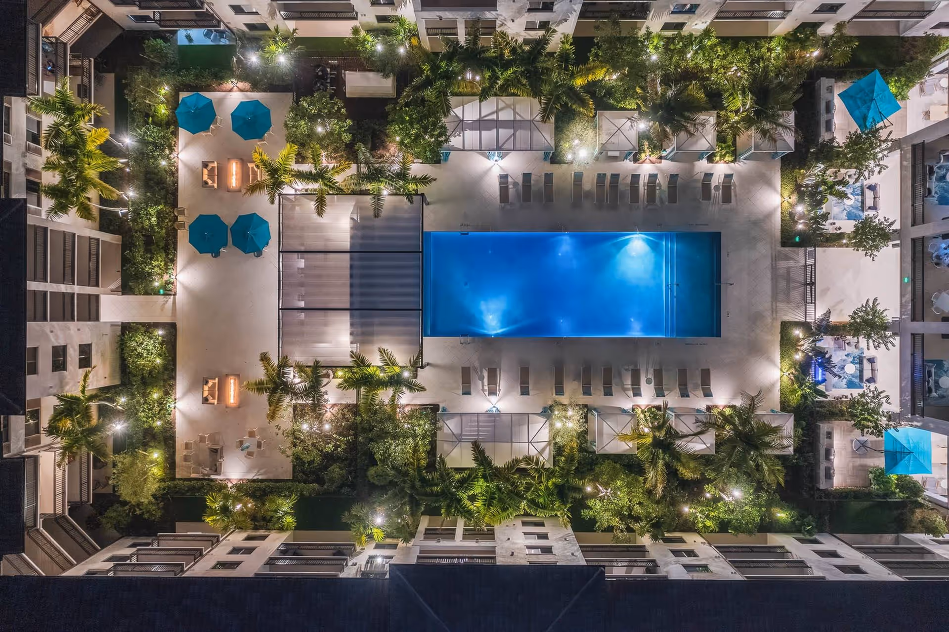 Aerial nighttime view of a residential complex courtyard featuring a large rectangular swimming pool illuminated with blue lights. Surrounding the pool are lounge chairs, palm trees, and several seating areas with tables and blue umbrellas. The area is well-lit with ambient lighting and greenery, creating a serene and inviting atmosphere.
