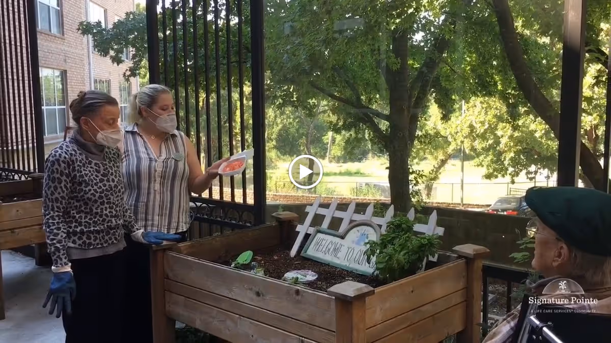 Two women wearing face masks and gloves stand next to a raised garden bed with plants and a small white picket fence decoration that says 'Welcome to our garden.' An elderly person in a wheelchair wearing a green cap watches them. The scene is outdoors with trees and a building in the background.