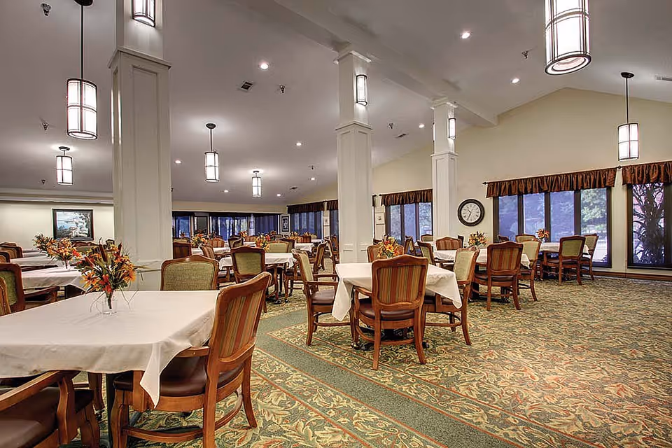 A spacious dining room with multiple tables covered in white tablecloths, each surrounded by wooden chairs with cushioned seats and backs. The room features large windows with brown valances, patterned carpet, and several hanging pendant lights providing warm illumination. Floral centerpieces decorate each table, and a clock is visible on the far wall.