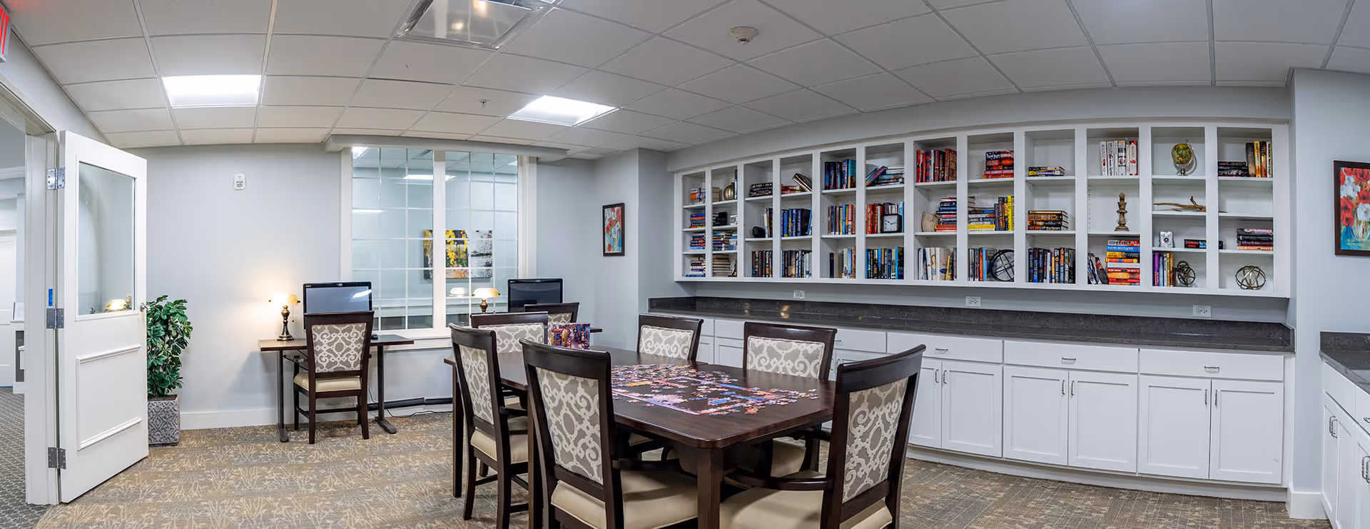 A well-lit room with a large wooden table surrounded by six upholstered chairs. The table has a puzzle in progress on it. Along one wall, there are built-in white shelves filled with books and decorative items, with cabinets below. Two computer desks with chairs and desktop computers are positioned against another wall. The room has a carpeted floor, light gray walls, and a white ceiling with recessed lighting. A potted plant is near an open door on the left side.