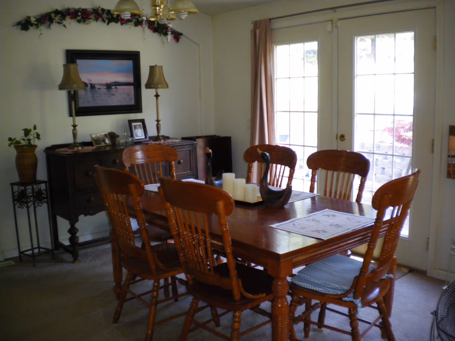 A dining room with a wooden table and six matching wooden chairs, two decorative black swan figurines and white candles on the table. Behind the table is a dark wooden sideboard with two lamps, framed photos, and a painting of a sunset over water on the wall. French doors with glass panes and beige curtains lead outside.