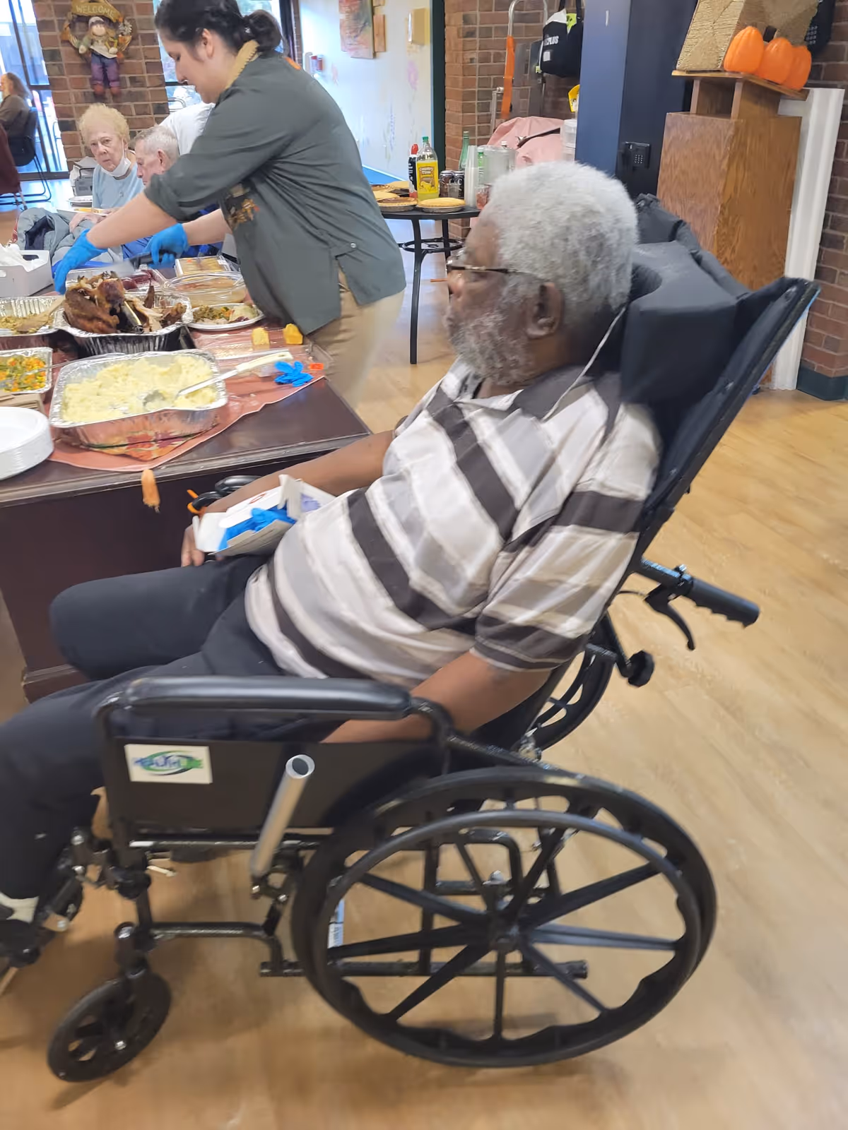 An elderly man in a wheelchair wearing a striped shirt is seated near a table with food trays. A woman wearing gloves is serving food to other elderly individuals seated at the table in a communal indoor setting with wooden floors and brick walls.