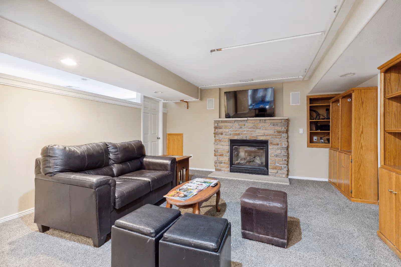 A cozy living room with a dark brown leather sofa, two black ottomans, and a small wooden coffee table with magazines and a remote control on it. The room features a stone fireplace with a mounted flat-screen TV above it, beige walls, carpeted floor, and wooden built-in shelves and cabinets on the right side.