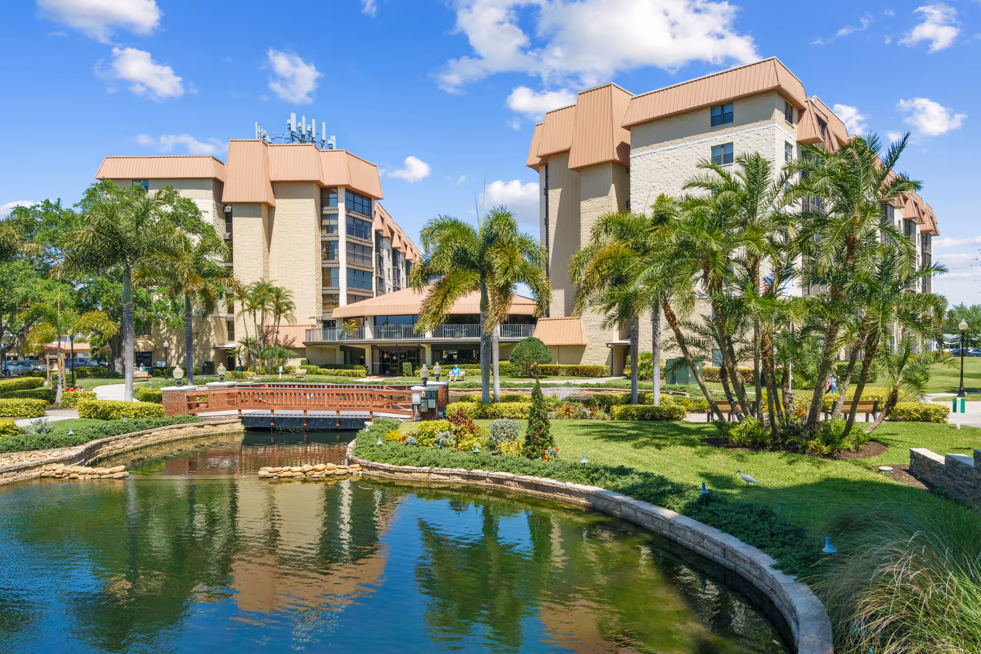 Exterior view of a senior living facility with beige buildings featuring brown roofs, surrounded by palm trees and well-maintained landscaping. In the foreground, there is a small pond with a curved stone edge and a wooden footbridge crossing over it under a bright blue sky with scattered clouds.