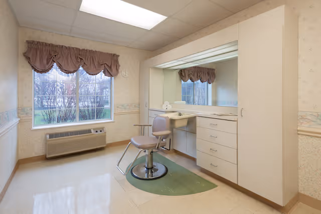 A small room with a single salon-style chair on a green mat in front of a large mirror and countertop with drawers. The room has a window with mauve valance curtains, a wall-mounted air conditioning unit below the window, and wallpaper with a floral border. The ceiling has fluorescent lighting.