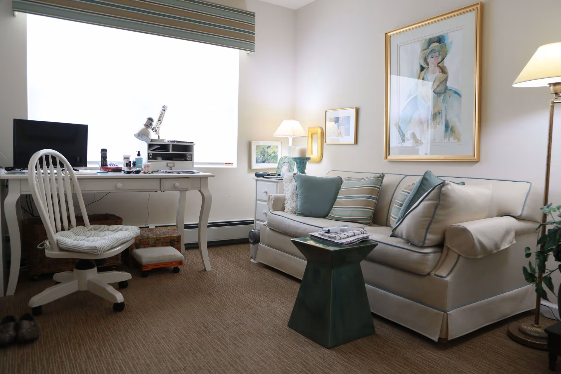 A cozy living room area with a beige sofa adorned with striped and solid pillows, a small green side table with folded towels on top, a white desk with a computer monitor, lamp, and office supplies, and a white swivel chair. The room has carpeted flooring, framed artwork on the walls, and a floor lamp in the corner.