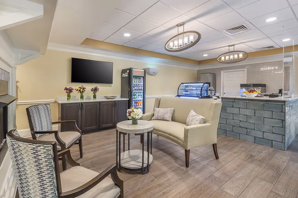 A cozy common area in a senior living facility featuring a small seating arrangement with two patterned armchairs and a cushioned loveseat around a round coffee table with a flower vase. Behind the seating area is a counter with a refrigerated display case, a Coca-Cola beverage cooler, and a cabinet with three flower vases. The room has warm lighting with two modern ceiling light fixtures and wood-style flooring.